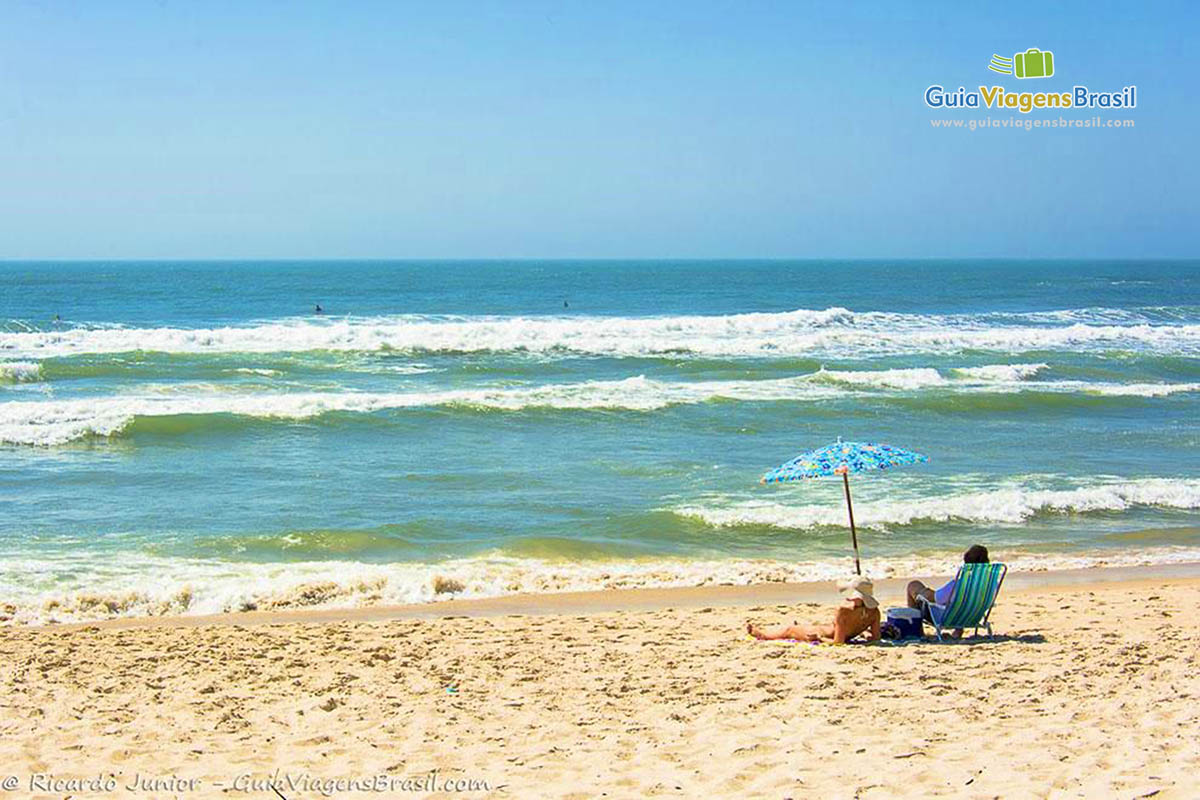 Imagem de duas pessoas sentadas em cadeiras de praia em baixo do guarda sol, olhando para o mar.