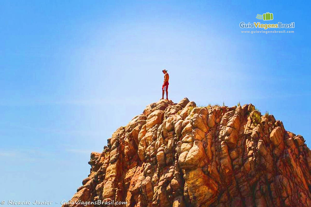 Imagem  de uma pessoa em cima de uma grande pedra avermelhada na praia.