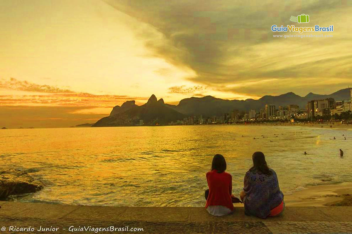 Imagem de duas amigas vendo o sol se pondo no horizonte na bela Praia Arpoador.