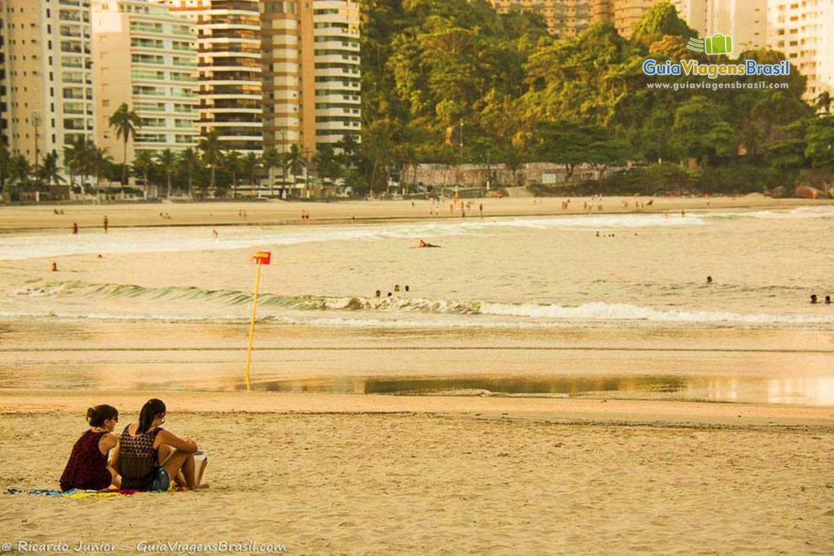 Imagem de duas amigas sentadas na areia conversando e admirando a paisagem.