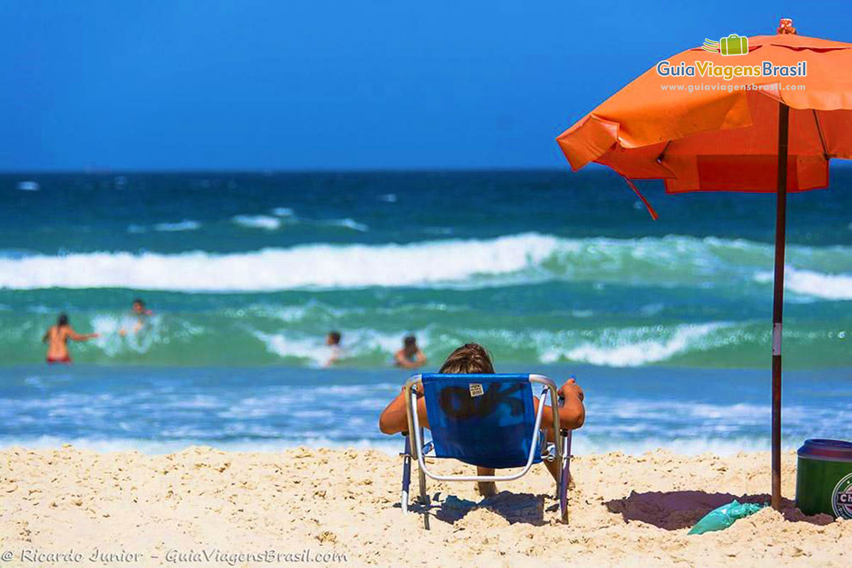 Imagem de um pessoa deitada na cadeira de praia com guarda sol ao lado, admirando a bela praia.