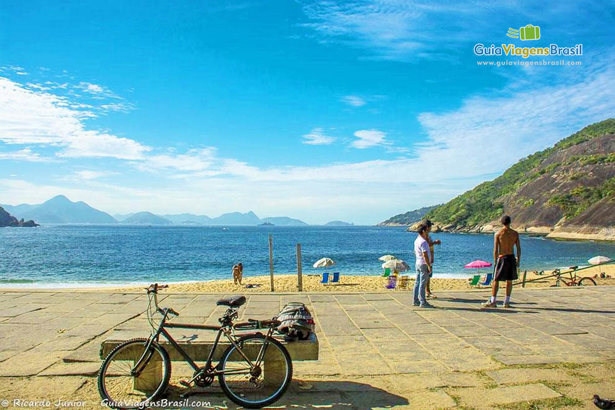 Imagem de uma bicicleta na orla e pessoas admirando a Praia Vermelha.