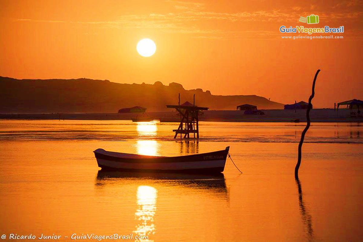 Imagem do céu bem alaranjado devido o sol e esta luz maravilhosa relfetindo o mar que possui um barco parado.
