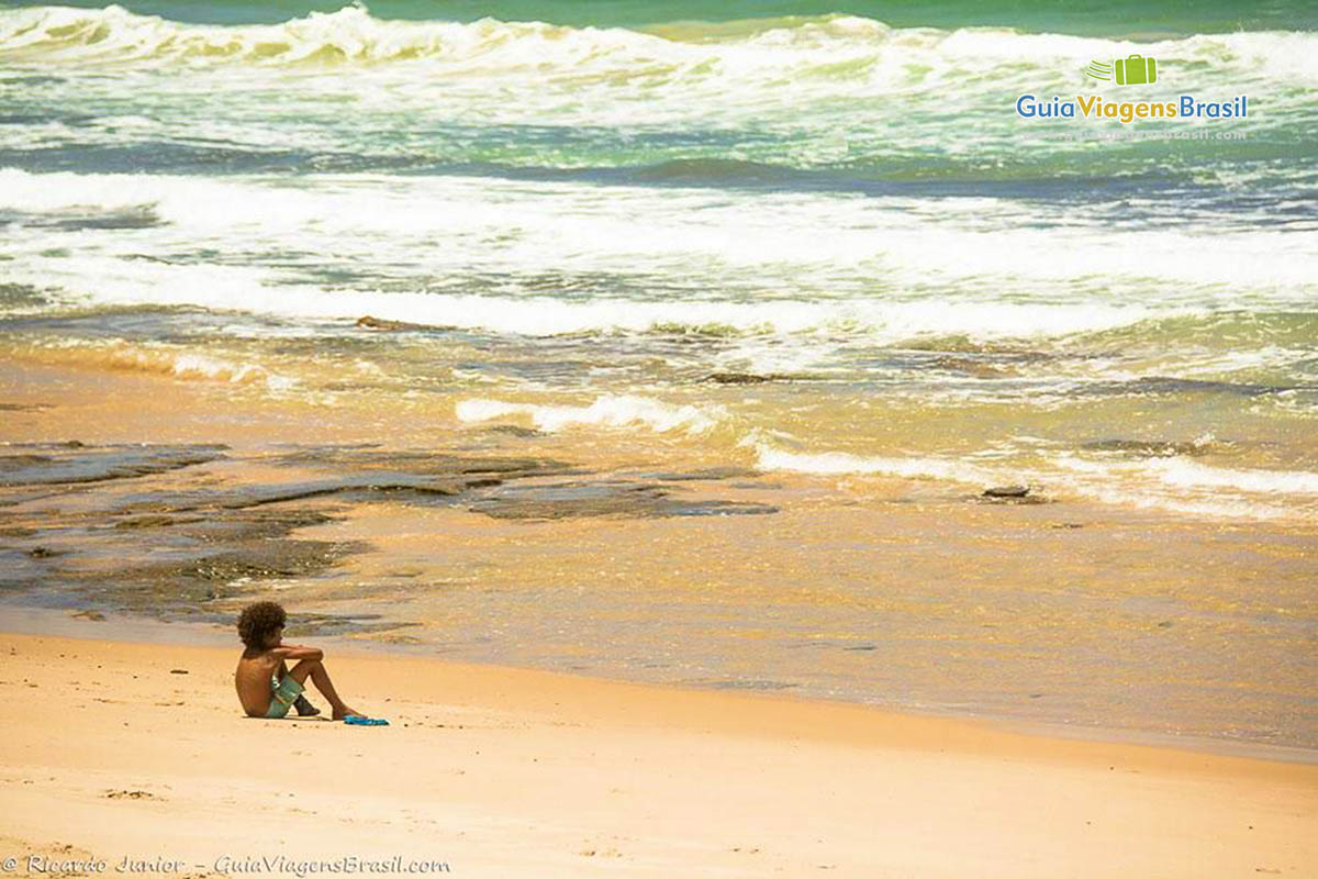 Imagem de uma criança sentado na areia da praia olhando para o mar.