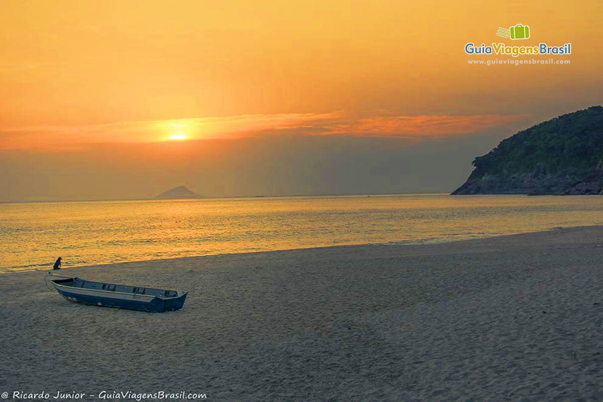 Imagem do sol se pondo no horizonte e um barco de pescador no mar, na Praia Santiago.
