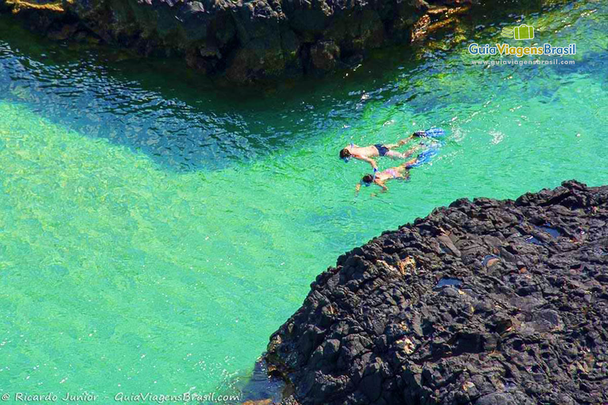 Imagem de dois turistas mergulhando entre duas pedras na Baía dos Porcos, em Fernando de Noronha, Pernambuco, Brasil.  