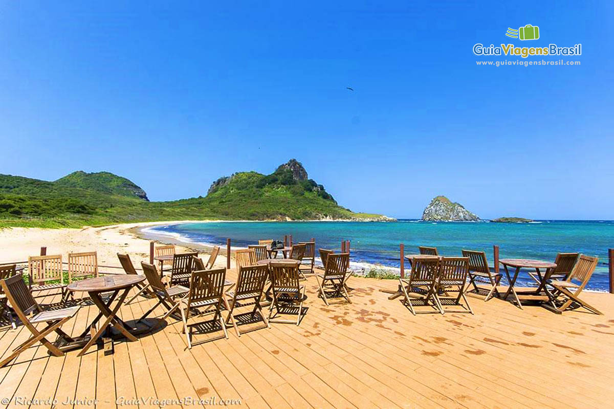 Imagem de cadeiras e mesas de madeira, colocadas em deck, e ao fundo vista da praia da Baía Sueste, em Fernando de Noronha, Pernambuco, Brasil.