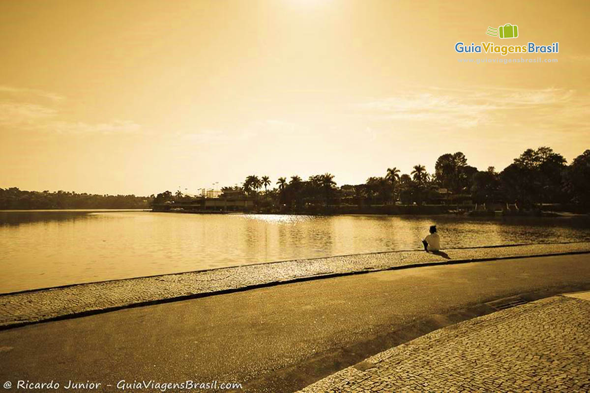 Imagem de uma pessoa sentada admirando o pôr do sol e a lagoa.