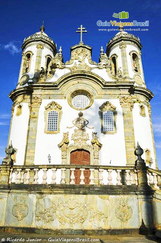Imagem do muro todo trabalhado com arte barroco mineira e a cima a igreja de São Francisco de Assis.