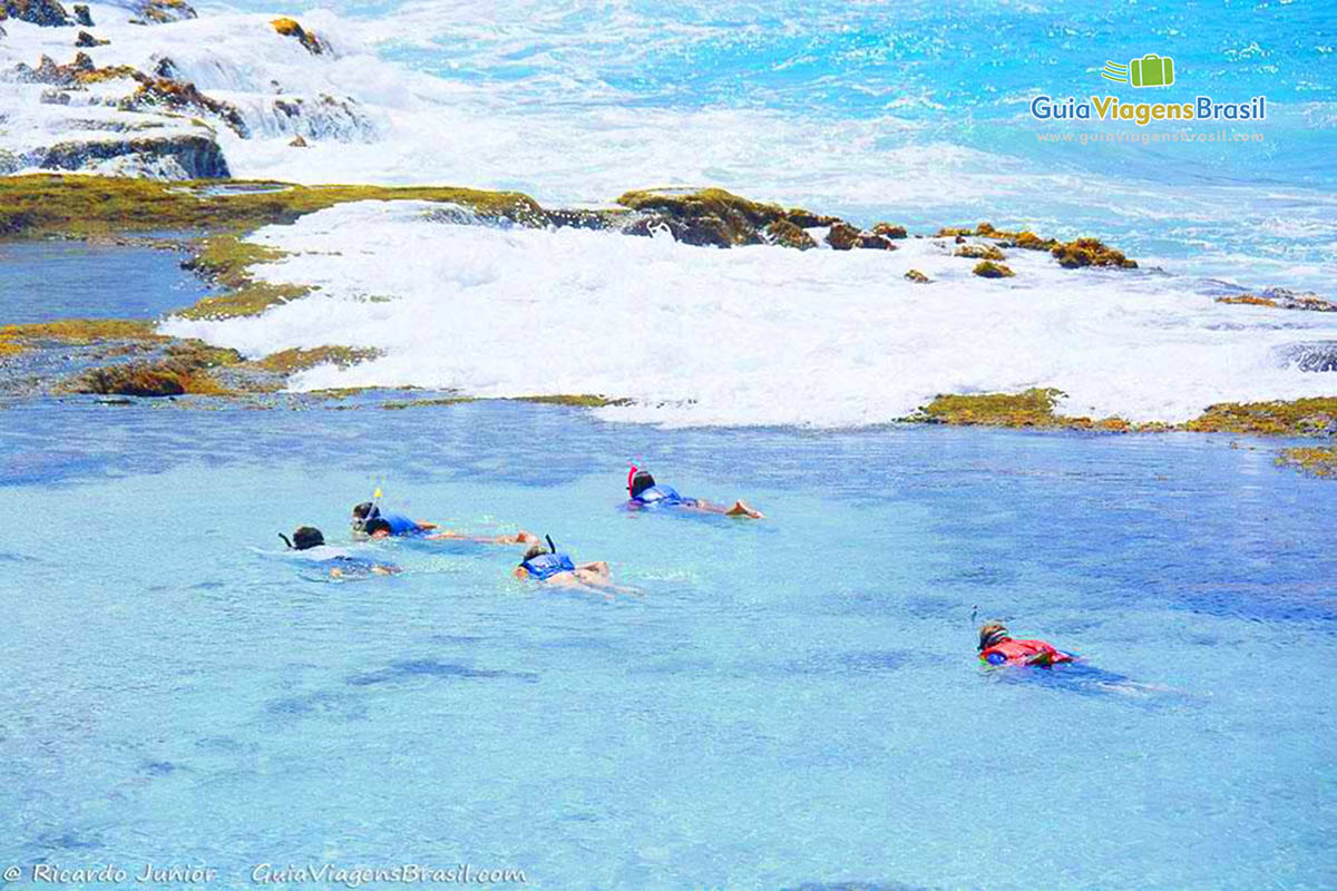 Imagem de turistas mergulhando de na piscina natural e atrás as águas azuis da Praia da Atalaia, em Fernando de Noronha, Pernambuco, Brasil.