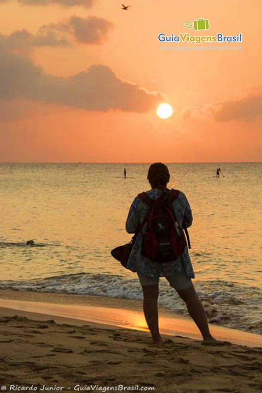 Imagem de turista admirando o pôr do sol e vendo pessoas praticar stand up, na Praia da Conceição, em Fernando de Noronha, Pernambuco, Brasil.