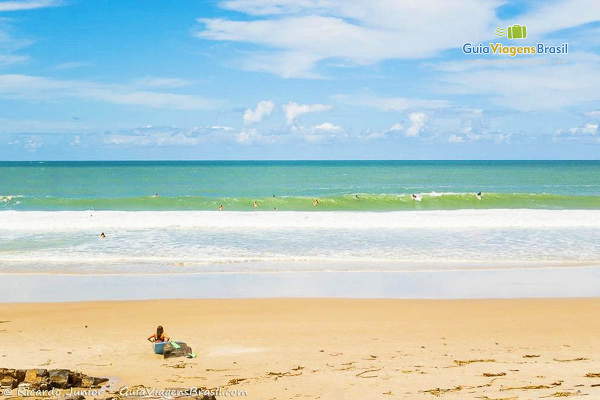 Imagem de uma surfista sentada na areia olhando os outros surfistas no mar.