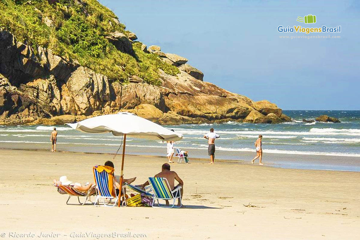 Imagem de turistas sentados na cadeira e com guarda sol, um aproveitando o sol para se bronzear e os outros admirando a Praia de Fora, da Ilha do Mel, Paraná, Brasil.