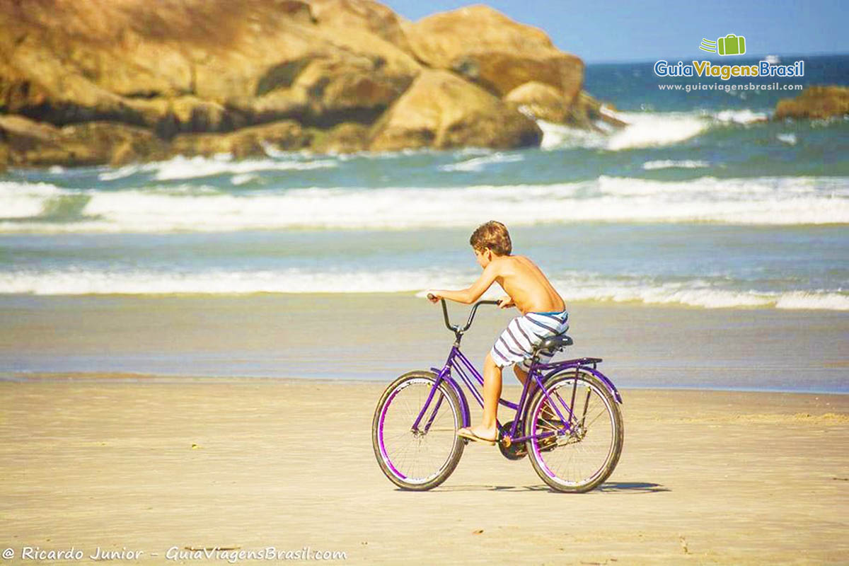 Imagem de menino andando de bicicleta na areia da Praia do Fora, Paraná, Brasil.