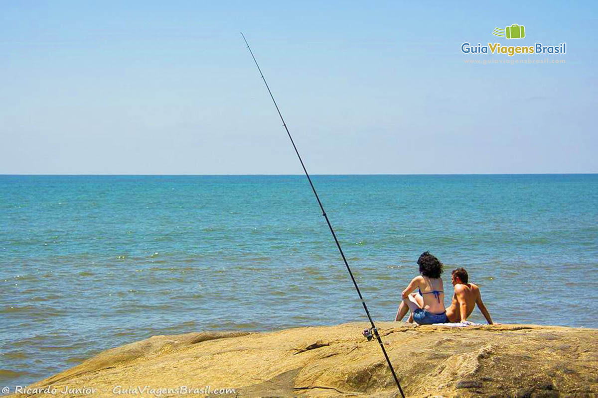 Imagem de vara de pescar presa nas pedras e casal admirando a paisagem, na Praia de Matinhos, em Santa Catarina, Brasil.
