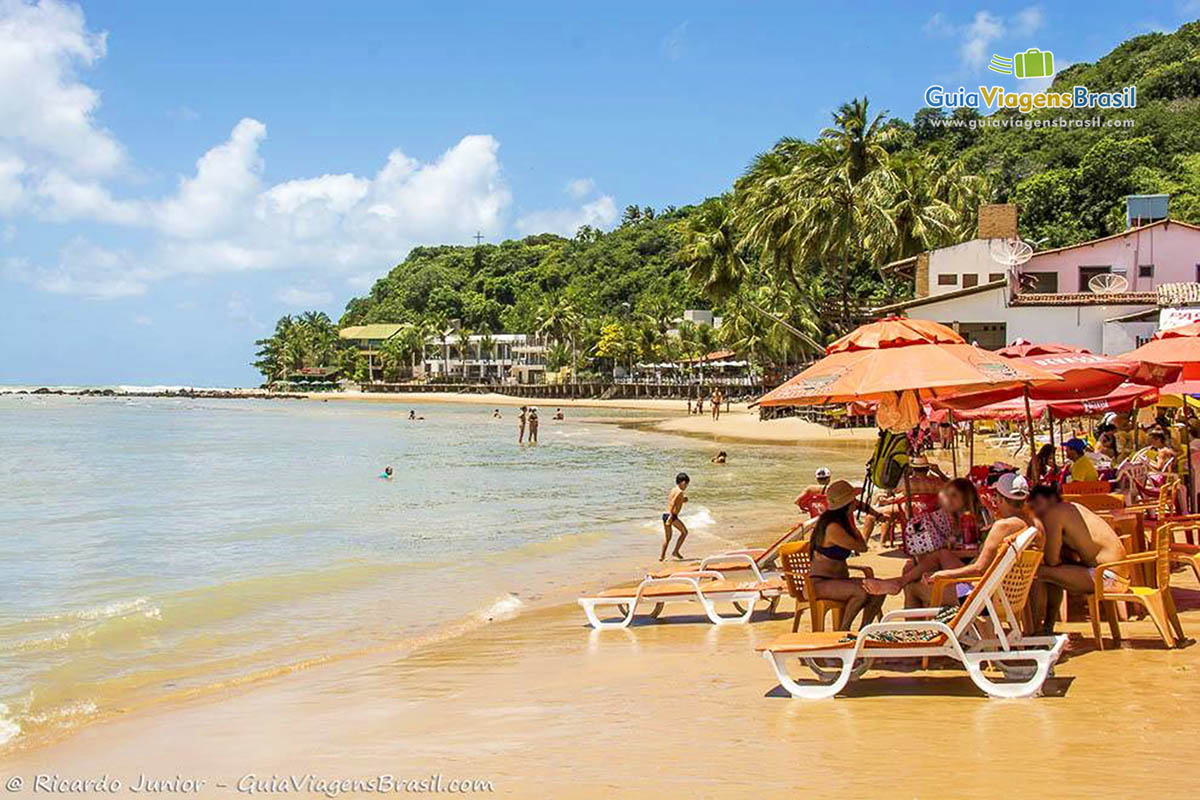Imagem de turistas com suas cadeiras e guarda sol na beira da praia.