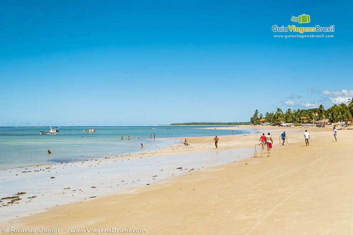 Imagem de turistas no mar e outros jogando bola nas areias da Praia de Tamandaré. 