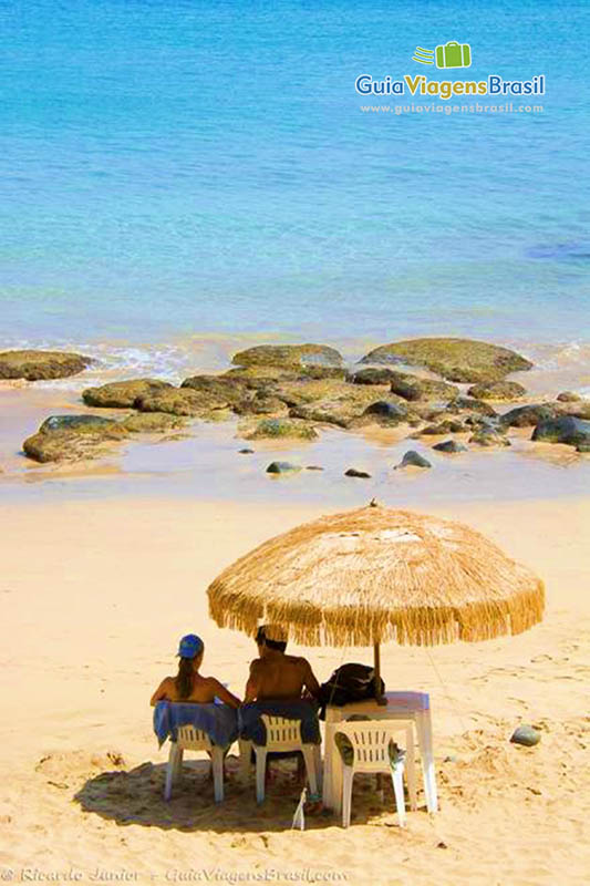 Imagem de casal sentados na cadeira na beira do mar com guarda sol de sapê, na Praia do Cachorro, em Fernando de Noronha, Pernambuco, Brasil.