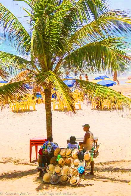 Imagem de um vendedor ambulante se protegendo do sol em baixo de um coqueiro.