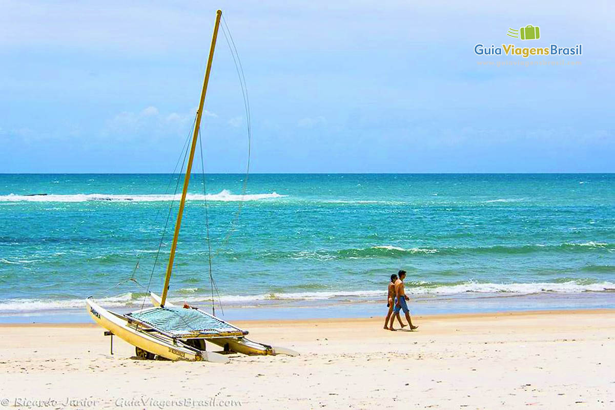 Imagem de barco na areia e casal caminhando ao fundo na beira da água, na Praia do Frances, em Maceió, Alagoas, Brasil.