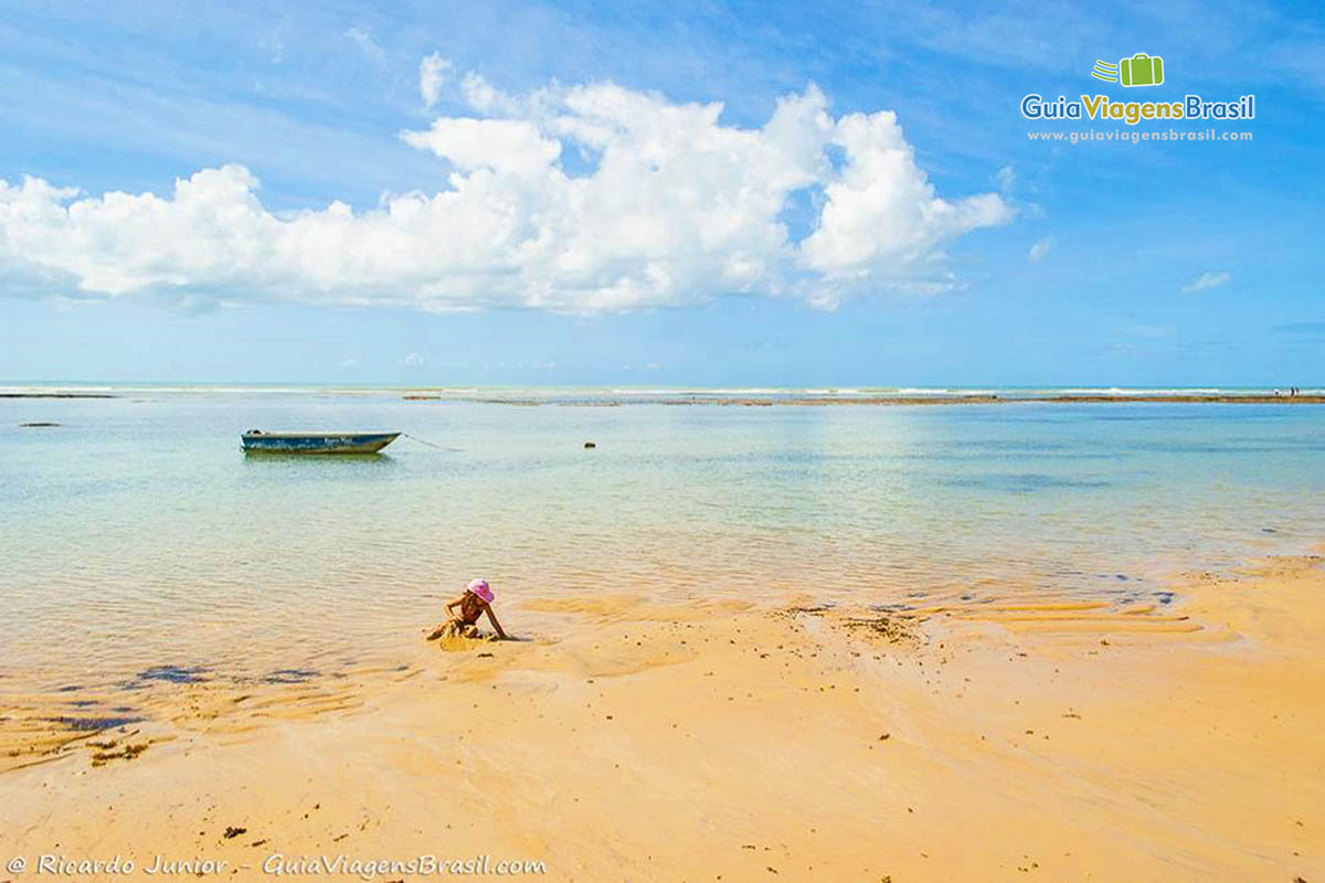 Imagem de criança brincando nas areias e ao fundo um barco de pescador na praia.