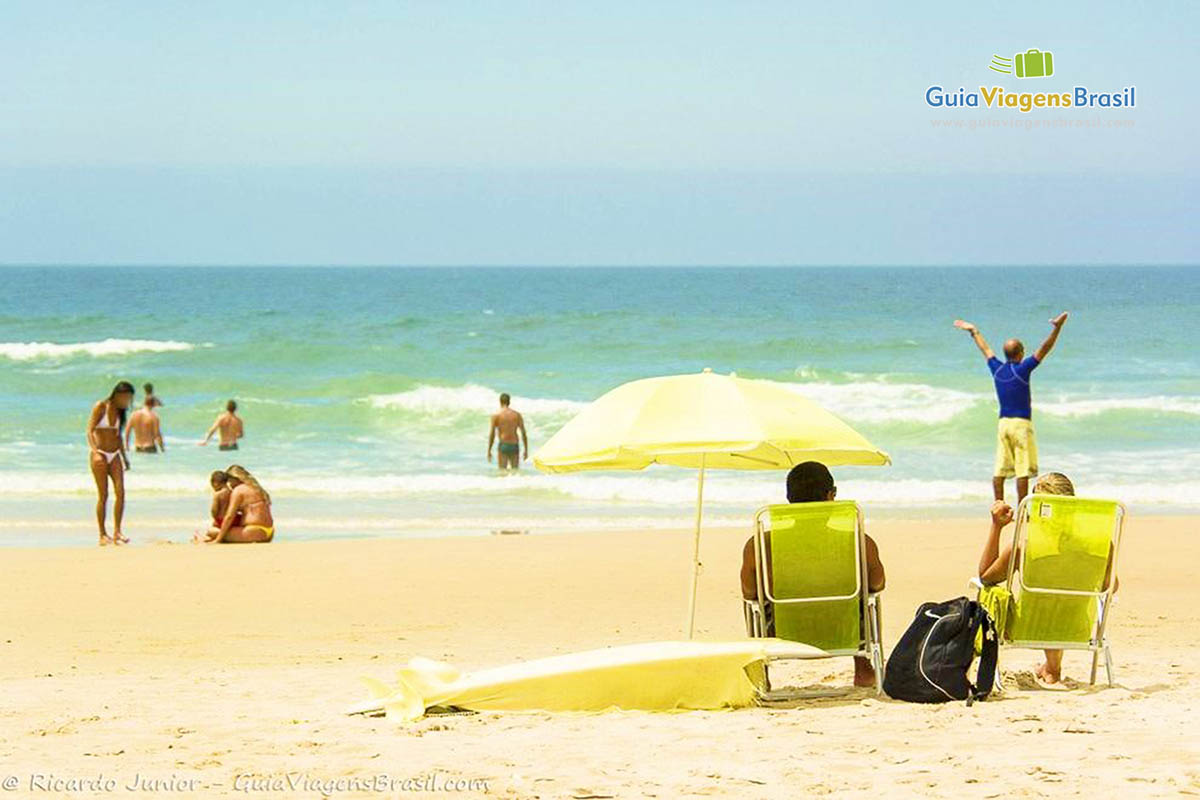 Imagem de uma pessoa sentado na cadeira de praia e outra na beira do mar sinalizando para alguém que está na água.