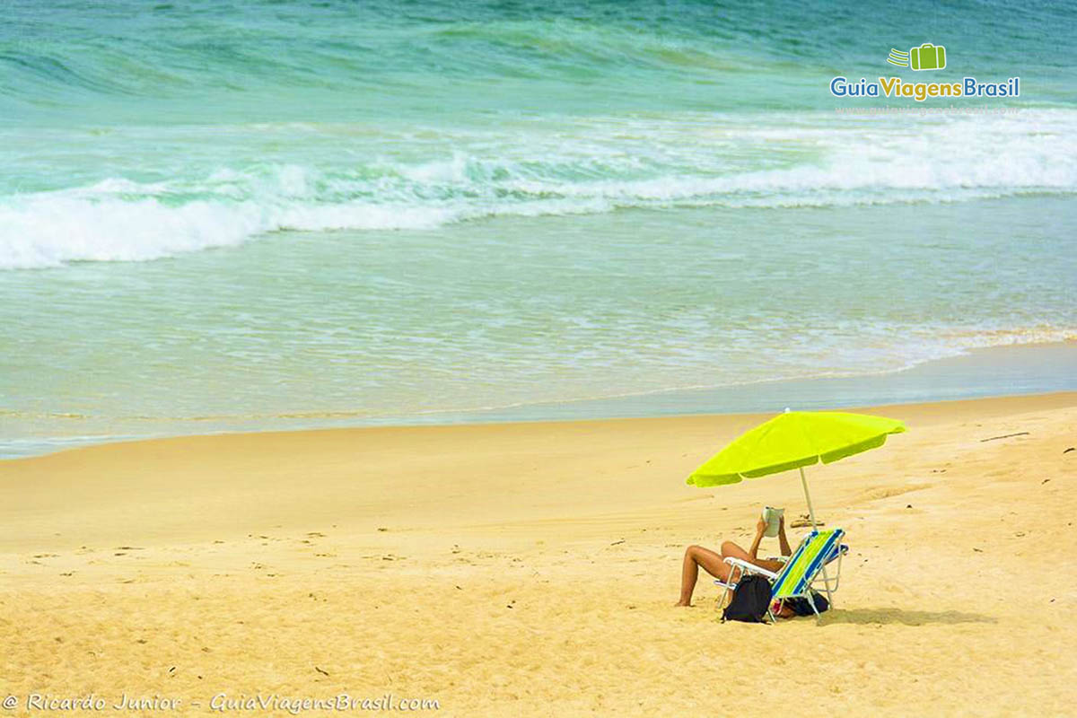 Imagem de uma pessoa lendo embaixo guarda sol verde limão e ao fundo o belo mar azul.
