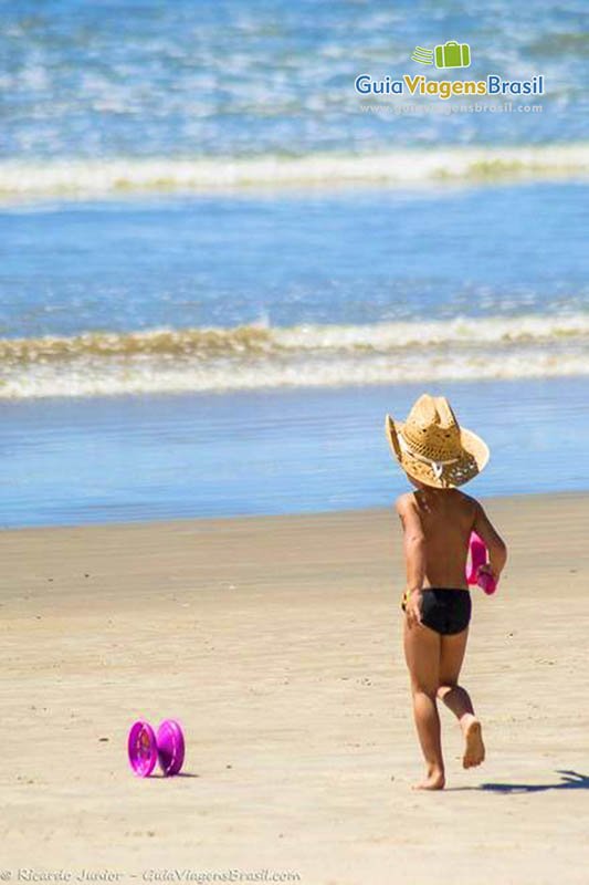 Imagem de um menininho andando nas beira da praia de chapéu.