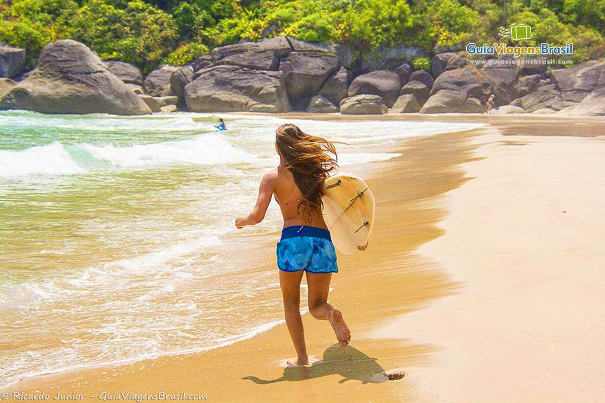 Imagem de menina correndo com sua prancha na beira da praia.