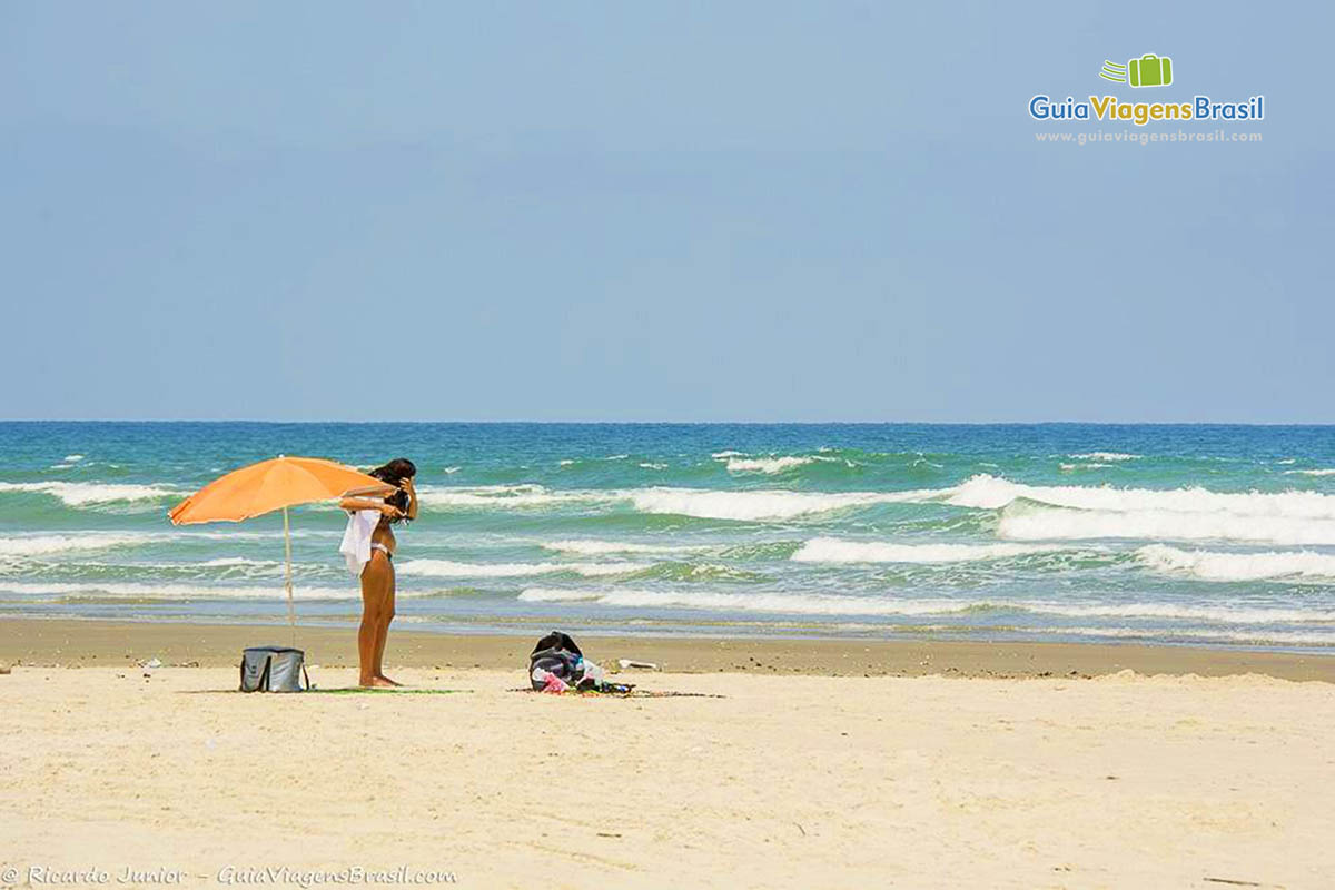 Imagem de menina em pé ao lado do guarda sol se arrumando na bela praia.