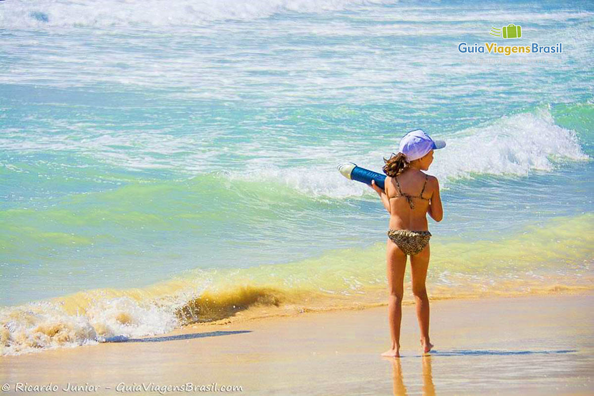 Imagem de uma menina em pé na beira do mar, brincando com uma arminha de água. 