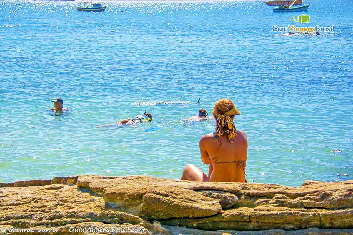 Imagem de uma moça sentada nas pedra olhando para mar e duas pessoas mergulhando com snorkel.