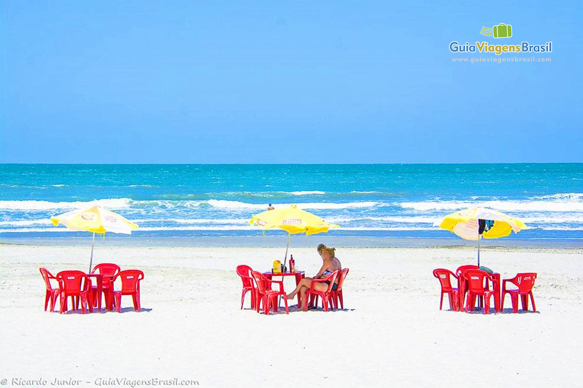 Imagem de mesas e cadeiras nas areias preparadas para receber os frequentadores da bela praia.