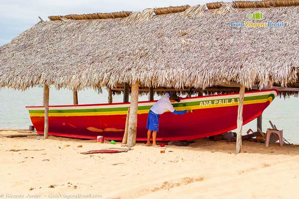 Imagem de um barco em baixo de barraca de sapê e uma pessoa pintando o barco.