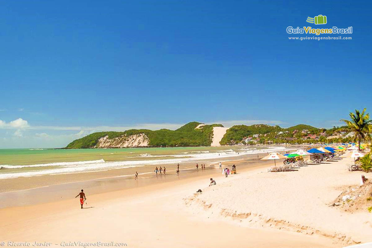 Imagem de turistas andando nas areias claras da linda Praia Ponta Negra.