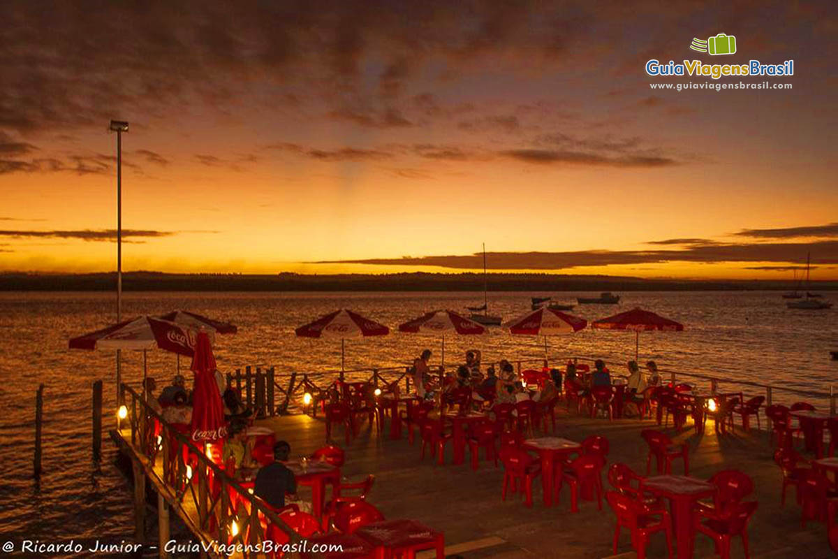 Imagem de cadeiras e mesas no deck iluminado e com o sol já encoberto no horizonte. Um belo fim de tarde na Praia Jacaré.