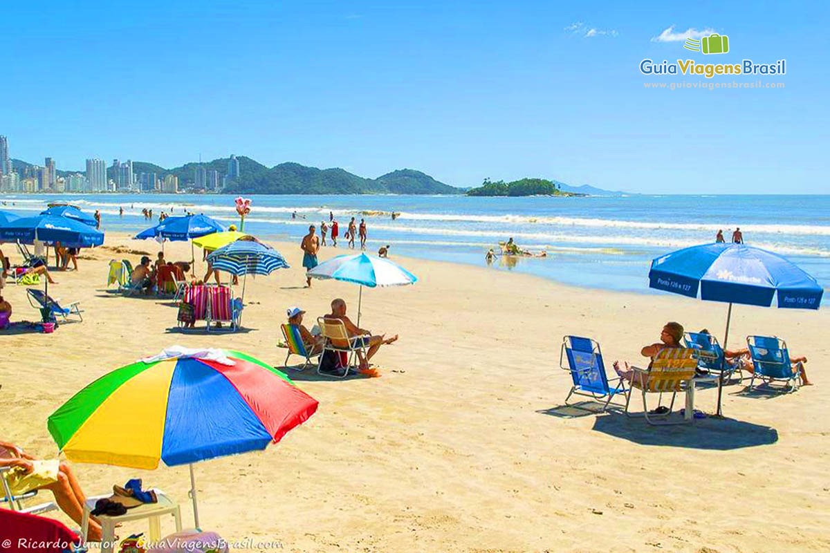 Imagem de turistas com guarda sol coloridos nas areias da praia.
