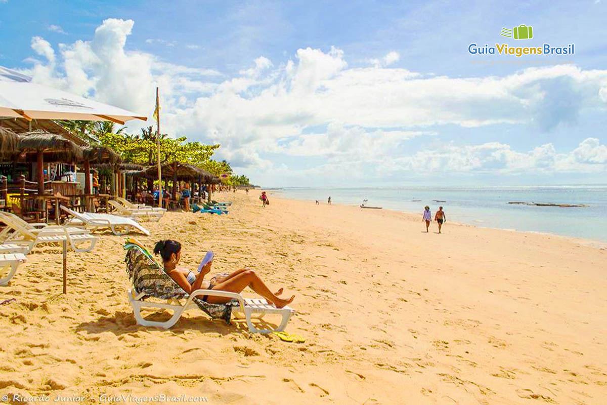 Imagem de turista tomando sol em uma cadeira e pessoas caminhando nas areias.