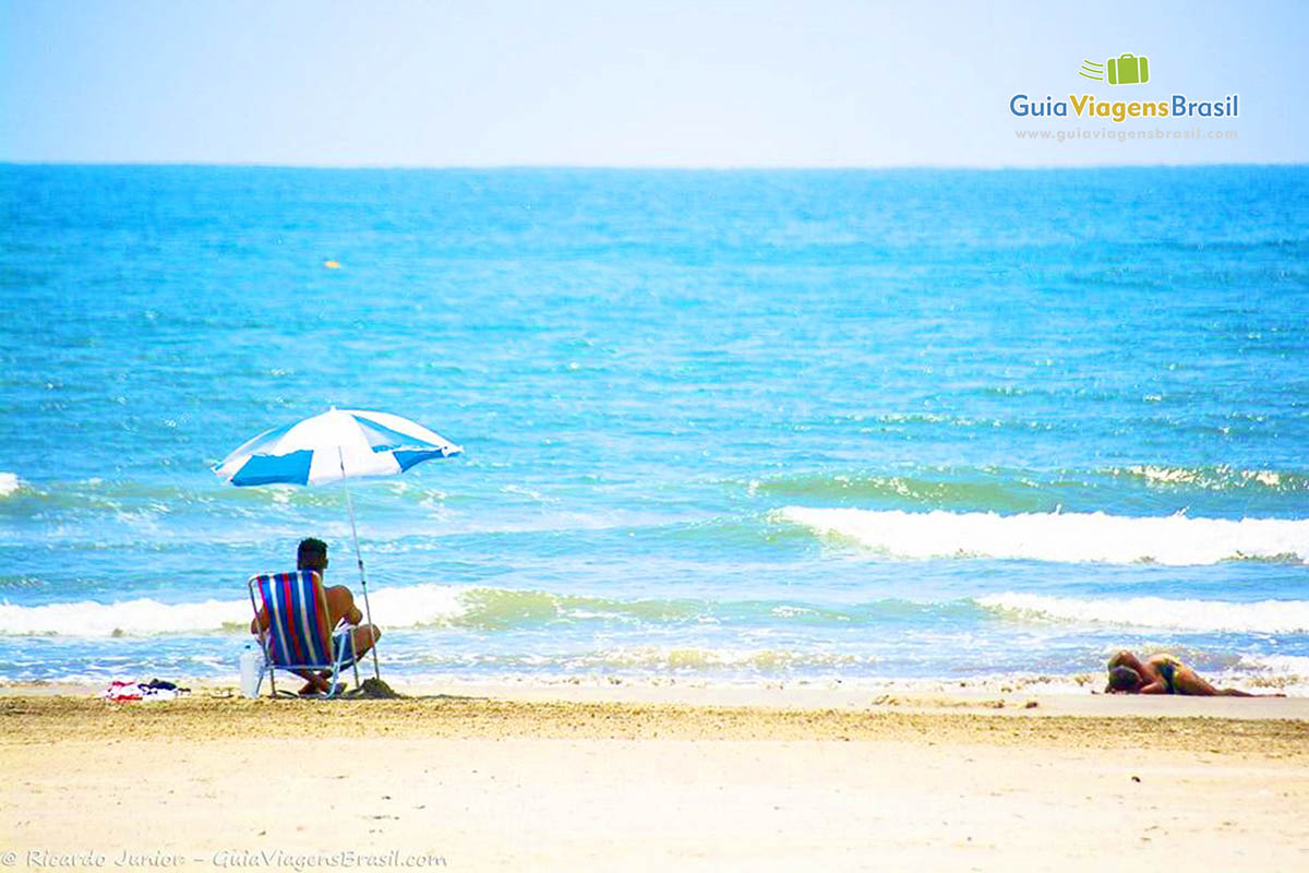 Imagem de um rapaz sentado embaixo de guarda sol admirando o mar da linda Praia Enseada.