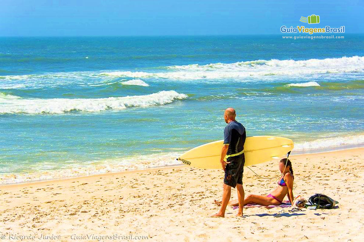 Imagem de surfista em pé olhando para mar com prancha na mão e uma moça sentada ao lado na areia.