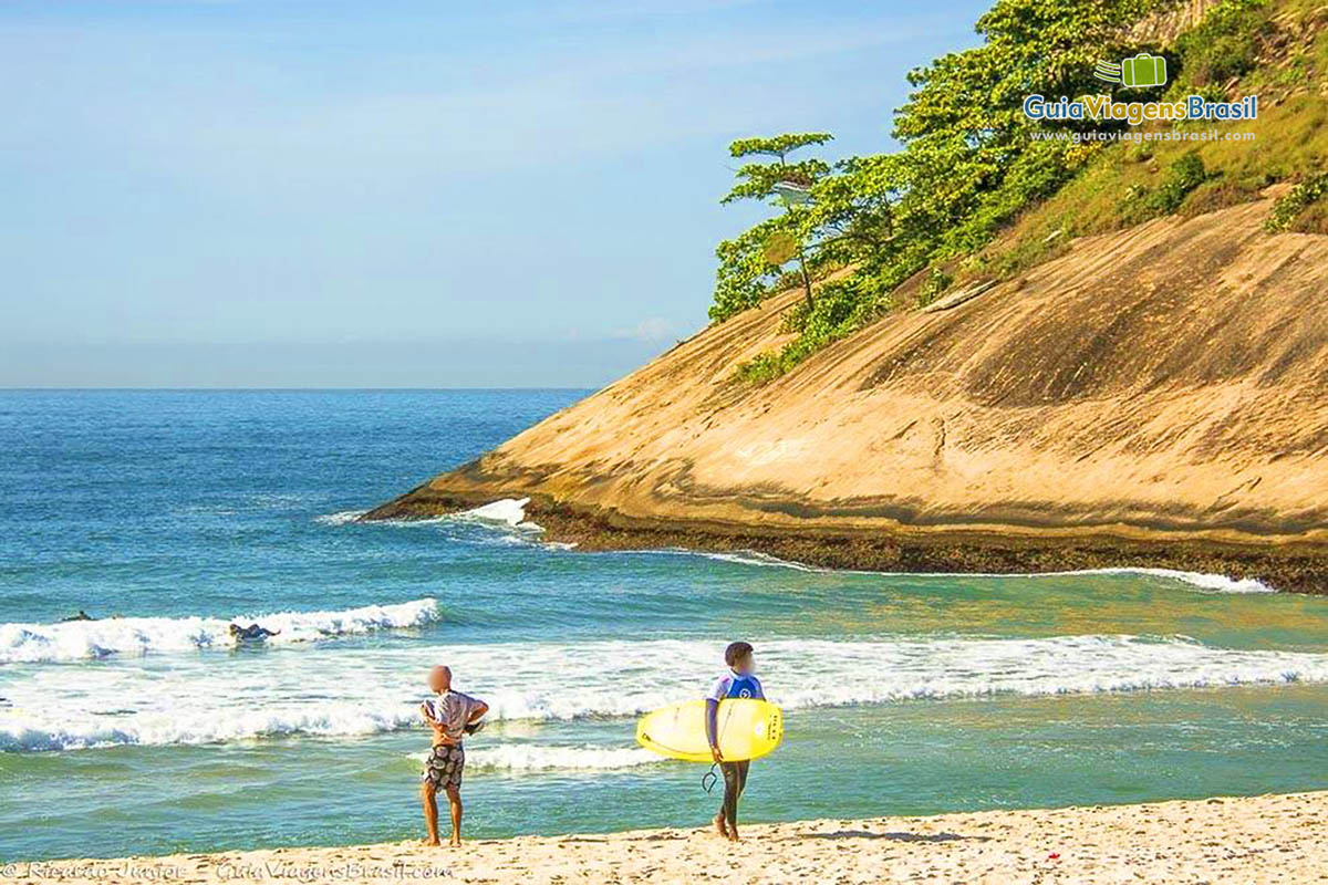 Imagem de surfista com prancha na mão na beira da praia.