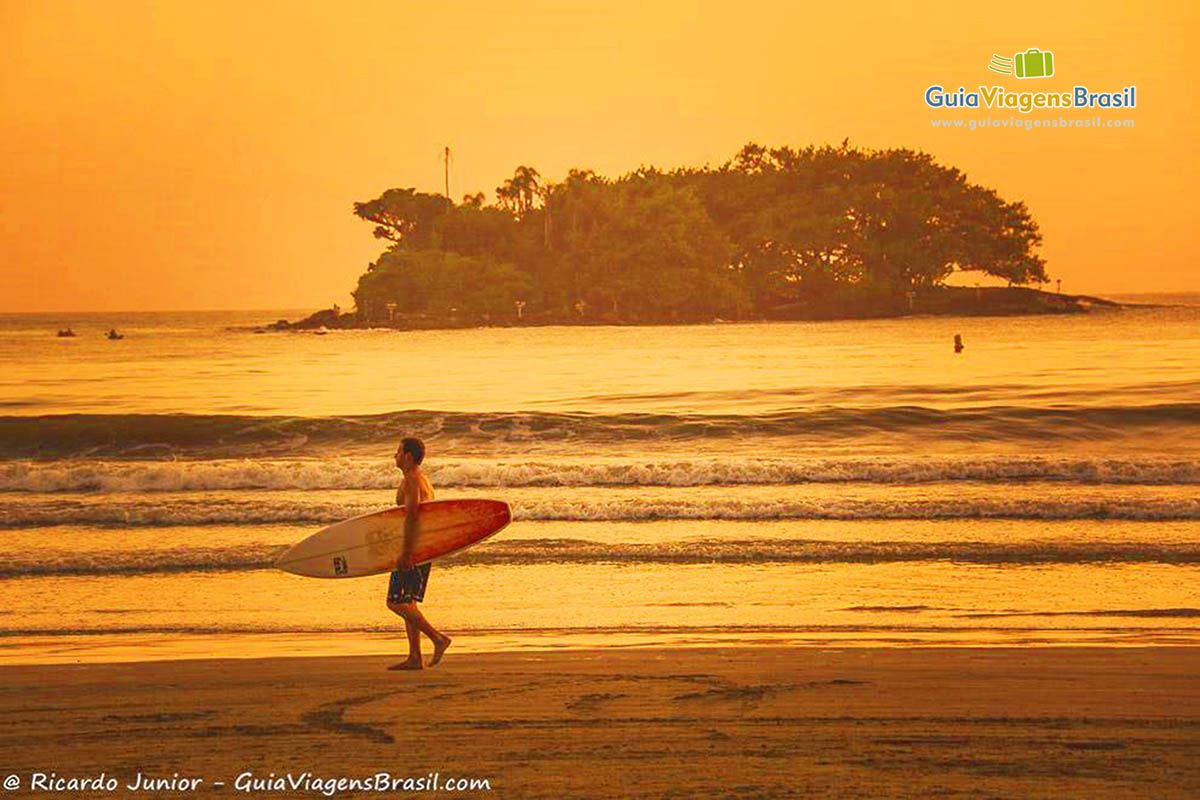 Imagem de surfista andando com prancha na beira do mar em um lindo fim de tarde, com céu alaranjado.